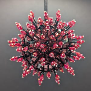 May include: A spherical, decorative ornament with a spiky design. The ornament is covered in red, faceted beads and crystals, with black wire accents. A black string is attached for hanging. The overall effect is a sparkling, festive decoration.