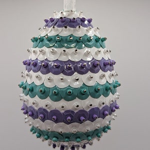May include: A white, purple, and teal egg-shaped ornament covered in sequins. The ornament has a white ribbon attached to the top.