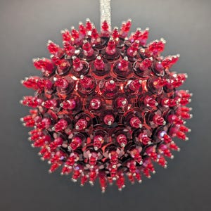 May include: A round, red Christmas ornament with a spiky, textured surface. The ornament is covered in small, red sequins and topped with red, pointed beads. A silver ribbon is attached for hanging. The ornament is against a dark gray background.
