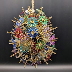 May include: A decorative sphere ornament with a multitude of colorful, flower-shaped elements. The flowers are in various colors, including red, blue, green, and purple, with gold-colored accents. A gold ribbon is attached for hanging.