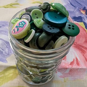 May include: A clear glass jar filled with a variety of green buttons. The buttons are different shapes, sizes, and shades of green, including olive, teal, and lime. Some buttons have stripes or floral patterns.