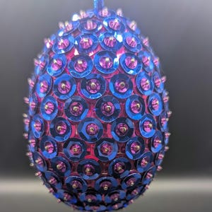 May include: An egg-shaped ornament covered in overlapping metallic blue sequins, each centered with a small, bright pink bead. The ornament is suspended by a thin blue cord attached to the top, creating a textured, reflective surface.