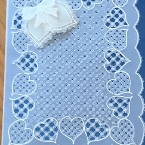 Puede incluir: Una tarjeta de papel blanco con fondo azul. La tarjeta tiene un diseño de encaje blanco con un patrón de corazón alrededor del borde. Un lazo de papel blanco está en la esquina superior izquierda.