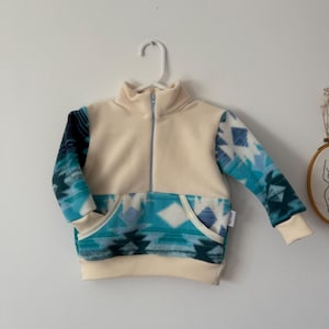 May include: A cream fleece pullover with a turquoise and blue geometric pattern on the sleeves and lower front. The pullover features a high collar, a front pocket, and a light blue zip. The garment is hanging on a white hanger.