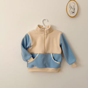 Può includere: Un pullover in pile a blocchi di colore crema e azzurro, con collo alto, cerniera frontale e tasca a marsupio. Le maniche e l'orlo hanno polsini color crema. L'indumento è appeso a una gruccia bianca.