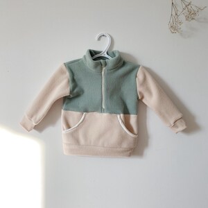 May include: A two-tone fleece pullover in sage green and beige, with a high collar and a front zip. The pullover has beige sleeves and a beige pocket. The garment is hanging on a white hanger against a white wall.