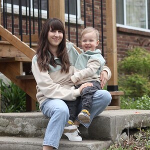 Peut inclure: Une femme et un enfant sont assis sur des marches en béton. Tous deux portent des sweat-shirts crème et vert menthe assortis et des jeans bleus. La femme porte des baskets blanches et l'enfant un pantalon gris et des chaussures à accents jaunes. Ils sourient.