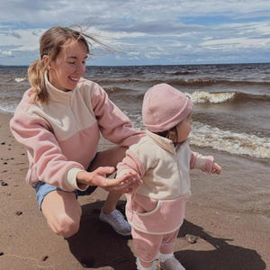 Op de afbeelding: Een vrouw en een peuter op een strand. Beiden dragen bijpassende roze en crèmekleurige sweatshirts en broeken. De peuter draagt ook een roze muts. De vrouw glimlacht en het kind kijkt naar het water. De zee is zichtbaar op de achtergrond.