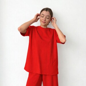 Può includere: Un completo composto da t-shirt e pantaloni rossi vivaci a gamba larga. La maglietta a maniche corte ha una vestibilità ampia e i pantaloni hanno un design fluido. L'outfit è realizzato in un tessuto morbido e tinta unita.