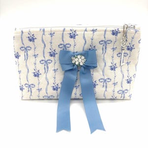 May include: A rectangular cosmetic bag with a floral and ribbon pattern in blue on a cream background. A light blue bow with a cluster of white flowers is attached to the front. A silver key charm hangs from the zipper.