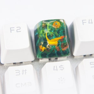 May include: A green and yellow resin keycap with a floral and fish design. The keycap is on a white keyboard.