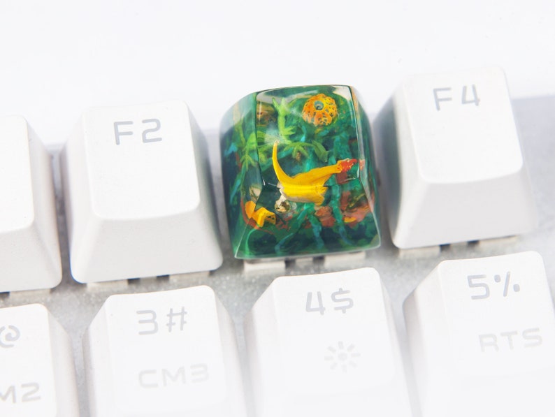 May include: A green and yellow resin keycap with a floral and fish design. The keycap is on a white keyboard.