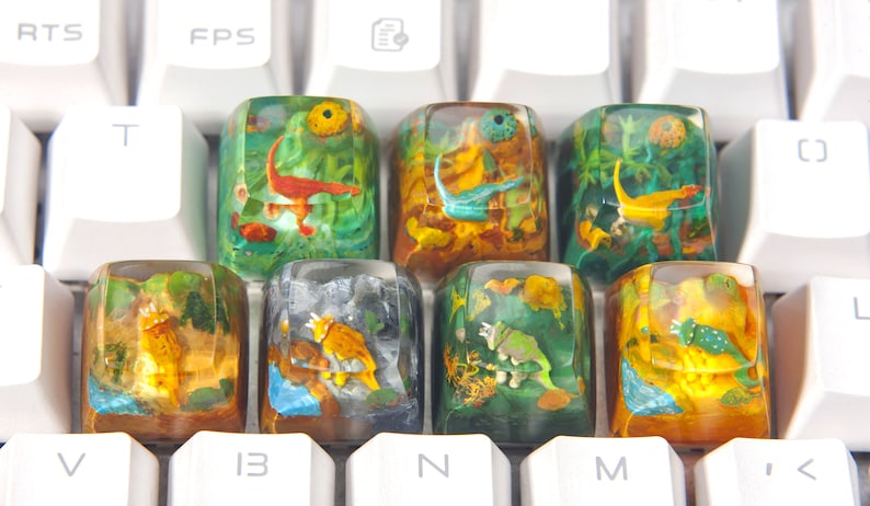 May include: Eight colorful keycaps with dinosaur and prehistoric animal illustrations encased in clear resin. The keycaps are arranged in two rows of four on a white keyboard.