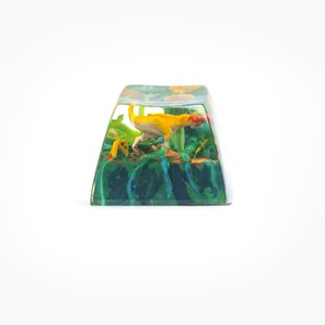 May include: A yellow and green resin keycap with a small dinosaur figurine inside. The dinosaur is in a crouching position with its tail curled up.