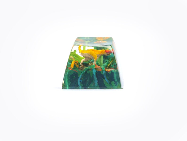 May include: A yellow and green resin keycap with a small dinosaur figurine inside. The dinosaur is in a crouching position with its tail curled up.