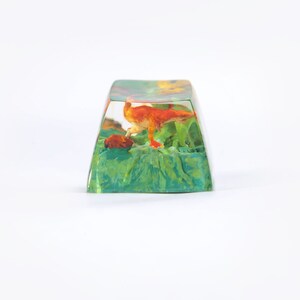May include: A clear resin keycap with a small orange dinosaur figurine inside. The dinosaur is standing on a green and brown base. The keycap is a popular item for keyboard enthusiasts.
