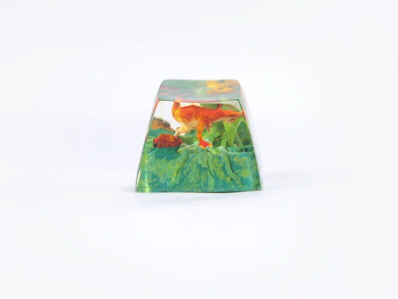 May include: A clear resin keycap with a small orange dinosaur figurine inside. The dinosaur is standing on a green and brown base. The keycap is a popular item for keyboard enthusiasts.