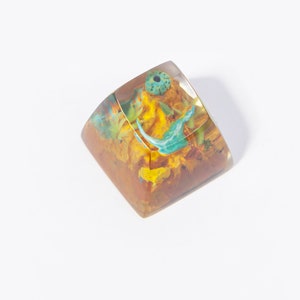 May include: A clear resin keycap with a yellow, orange, and blue floral design. The keycap is a square shape and has a small hole in the center.
