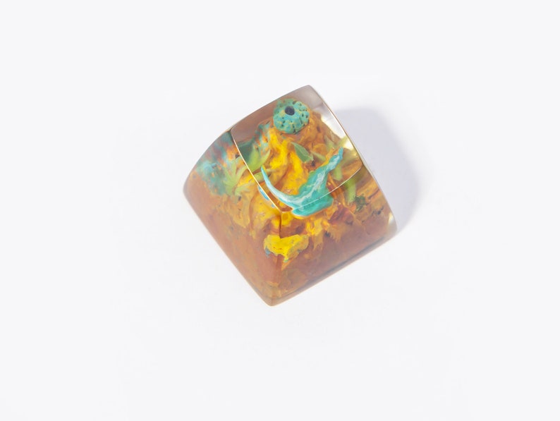 May include: A clear resin keycap with a yellow, orange, and blue floral design. The keycap is a square shape and has a small hole in the center.