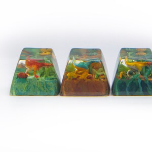 May include: Three resin keycaps with a dinosaur design. Each keycap features a different colored dinosaur in a green, brown, and blue landscape.