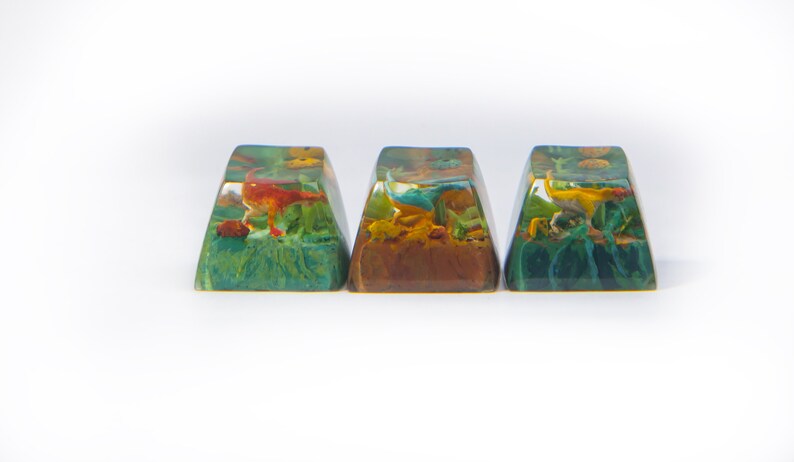 May include: Three resin keycaps with a dinosaur design. Each keycap features a different colored dinosaur in a green, brown, and blue landscape.