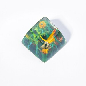 May include: A green, yellow, and orange resin keycap with a floral design. The keycap is shaped like a square with rounded corners.