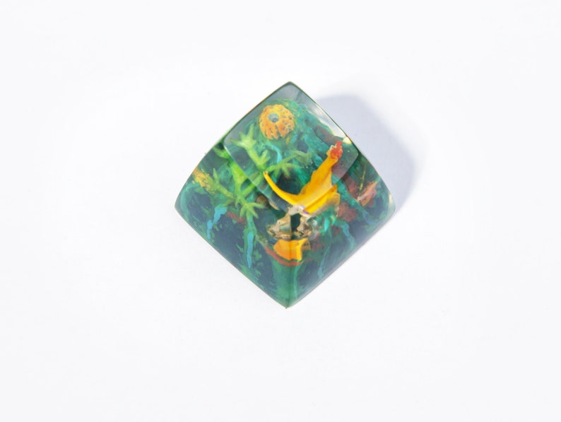 May include: A green, yellow, and orange resin keycap with a floral design. The keycap is shaped like a square with rounded corners.