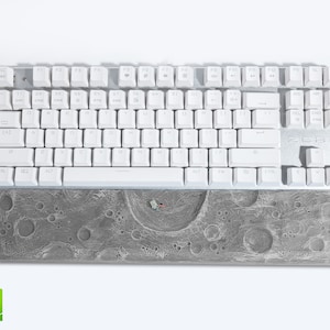 May include: A white mechanical keyboard with white keycaps and a silver frame. Below the keyboard is a gray wrist rest with a detailed moon surface design and a small astronaut figure.