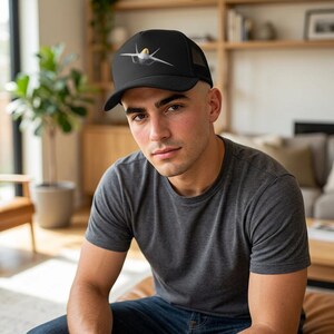 Può includere: Un cappellino trucker nero con la grafica di un jet da combattimento grigio sul pannello frontale. Il cappello presenta una corona strutturata, una visiera curva e una parte posteriore in rete. L'illustrazione del jet mostra un tettuccio giallo.