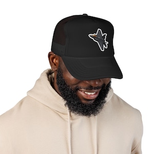 Peut inclure: Une casquette de camionneur noire avec un graphique représentant un avion de chasse gris foncé doté d'un cockpit orange. La casquette présente un panneau avant solide et un panneau arrière en maille, avec l'illustration centrée sur l'avant.