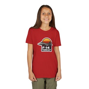 May include: A red t-shirt featuring a graphic of an F-14 Tomcat fighter jet against a sunset design. The text "F-14 TOMCAT" is below the jet. The shirt is being worn by a person.