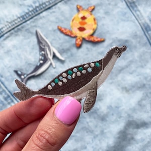 May include: An embroidered patch of a prehistoric marine reptile, brown and white with green and white spots. The patch is on a denim background.