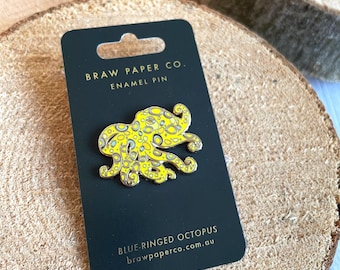 May include: A yellow and blue enamel pin shaped like a blue-ringed octopus, mounted on a black display card. The card features the text BRAW PAPER CO. ENAMEL PIN at the top and BLUE-RINGED OCTOPUS brawpaperco.com.au at the bottom.