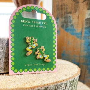 May include: A pair of enamel earrings shaped like green tree frogs with yellow, blue, and orange details, displayed on a pink and green card. The card reads "BRAW PAPER CO. ENAMEL EARRINGS" and "Green Tree Frogs".
