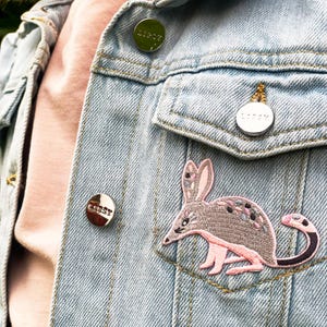 May include: A denim jacket with a pink and grey embroidered bandicoot patch, a silver pin with the word "LIBBY" on it, and two other silver pins with the word "LIBBY" on them.