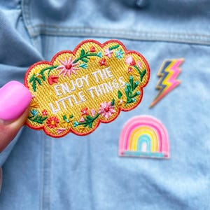 May include: Yellow embroidered patch in the shape of a cloud with floral details. The patch reads "Enjoy the little things".