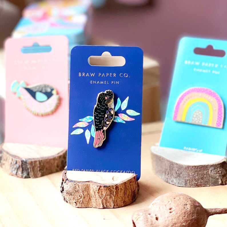 Pode incluir: Um cart&atilde;o azul e branco com o texto "Braw Paper Co. Enamel Pin" e um alfinete de esmalte preto e vermelho de um p&aacute;ssaro com o texto "Red-tailed Black Cockatoo".