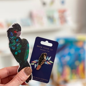 Pode incluir: Um broche de esmalte azul e dourado com um cacatua preto com uma crista rosa e amarela. O broche &eacute; embalado em um cart&atilde;o azul com o texto "Braw Paper Co. Enamel Pin" e "Wiled Black Cockatoo brawpaperco.com.au".
