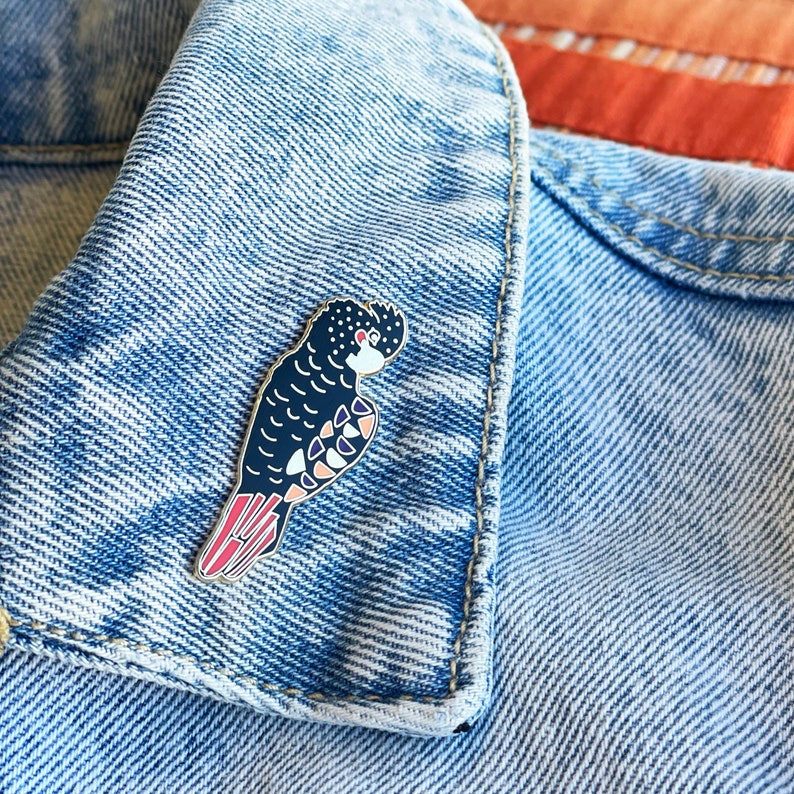 Pode incluir: Um broche de esmalte preto e branco de um cacatua com detalhes vermelhos, rosa e azuis. O broche est&aacute; em uma jaqueta jeans azul.