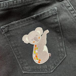 May include: A grey koala embroidered patch with orange and green accents on a black denim background.
