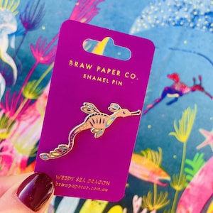 May include: A gold-toned enamel pin shaped like a weedy sea dragon with light orange, blue, and yellow accents. The pin is attached to a rectangular purple display card featuring the text BRAW PAPER CO. ENAMEL PIN and WEEDY SEA DRAGON.