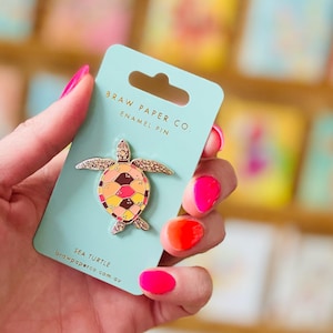 May include: A sea turtle enamel pin attached to a light blue backing card. The pin features a multi-coloured shell with shades of yellow, orange, pink, and dark red. The card displays the text BRAW PAPER CO. ENAMEL PIN and SEA TURTLE.