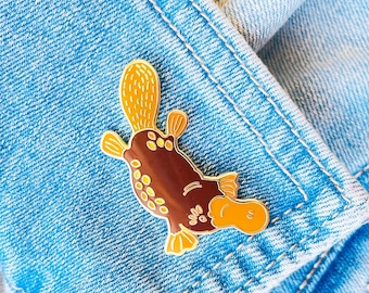 Pode incluir: Um broche de esmalte marrom e laranja em forma de ornitorrinco. O broche está sobre um fundo de ganga azul. O ornitorrinco tem cauda amarela, pés palmados e bico de pato.