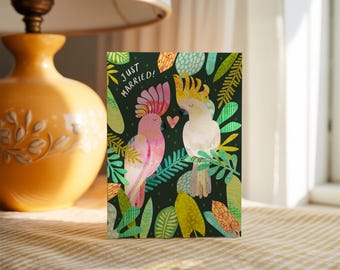 May include: A rectangular greeting card with a dark green background featuring two cockatoos, one pink and one white, perched among colorful tropical leaves. The text "JUST MARRIED!" is printed in white near the top left, with a small pink heart.