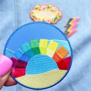 May include: A round, blue embroidered patch with a rainbow arc and a yellow beach scene. The rainbow is made up of 10 different coloured stripes.