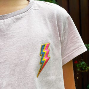 May include: A light pink t-shirt with a colourful lightning bolt embroidered on the front.