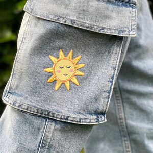 May include: A pair of blue denim cargo trousers with a yellow embroidered sun with a smiling face on the front pocket.