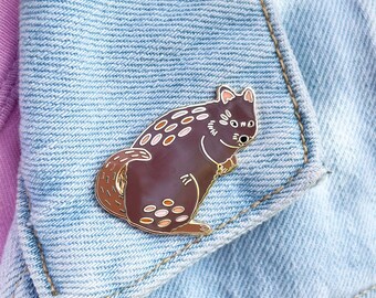 May include: A dark brown enamel pin shaped like a quokka with gold-colored metal outlines. The quokka features small, light-colored oval spots on its back and tail, with a rounded body, pointed ears, and a small snout, attached to light blue denim fabric.