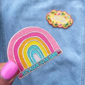 May include: An embroidered patch featuring a rainbow design with the text "It's cool to be kind" and a small pink heart. The patch is displayed on a light blue denim background.