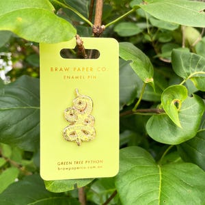 May include: A gold-toned enamel pin shaped like a coiled snake, displayed on a lime green rectangular backing card. The card features the text BRAW PAPER CO. ENAMEL PIN at the top and GREEN TREE PYTHON brawpaperco.com.au at the bottom.